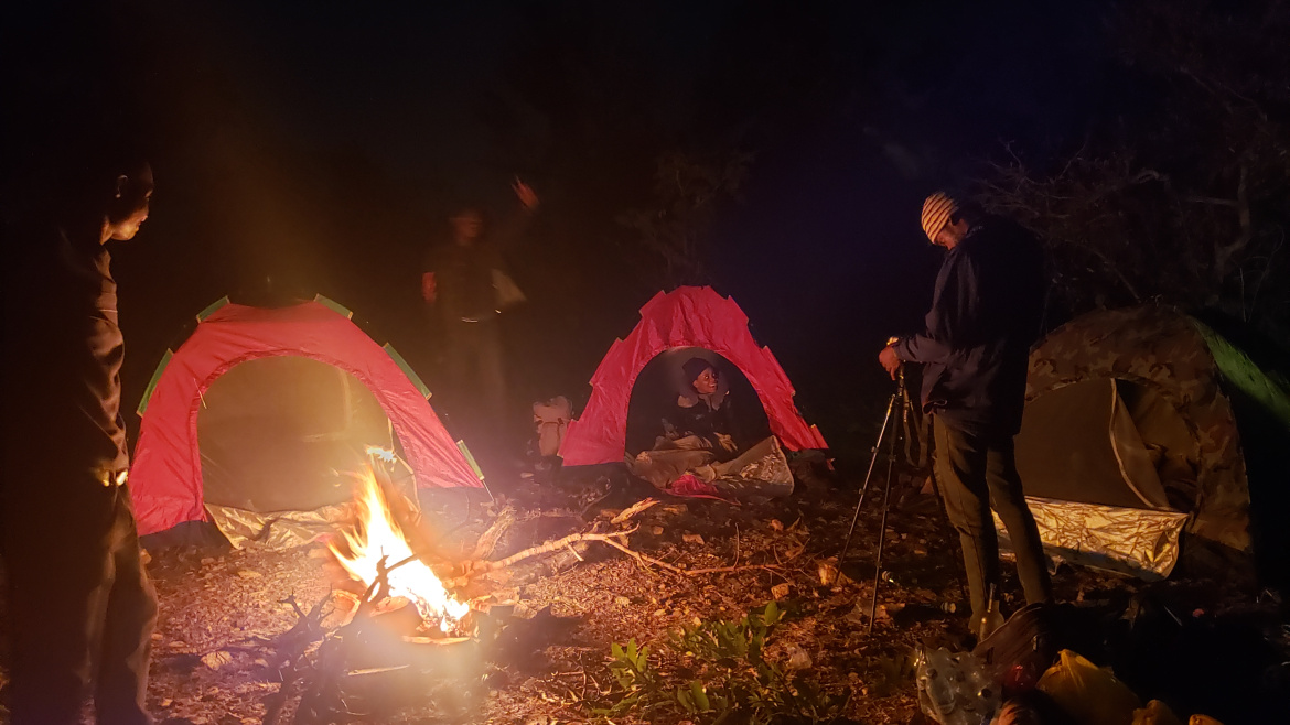 Campers gathered around a glowing bonfire at night, with tents lit up in the background during the first Crossover Camp — a moment of warmth, reflection, and togetherness under the open sky.”