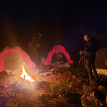 Campers gathered around a glowing bonfire at night, with tents lit up in the background during the first Crossover Camp — a moment of warmth, reflection, and togetherness under the open sky.”